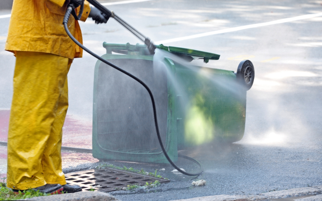 Worker in protective gear pressure washing a green wheelie bin